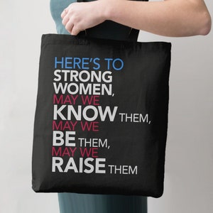 May include: Black tote bag with the text "HERE'S TO STRONG WOMEN, MAY WE KNOW THEM, MAY WE BE THEM, MAY WE RAISE THEM" in blue, pink, and white.