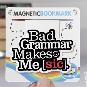 May include: A white magnetic bookmark with a rainbow design and the text "Bad Grammar Makes Me [sic]" in black. The bookmark is held by a hand wearing a beige knitted sweater. The text "BootsTEES MAGNETIC BOOKMARKS" is printed on the bookmark.