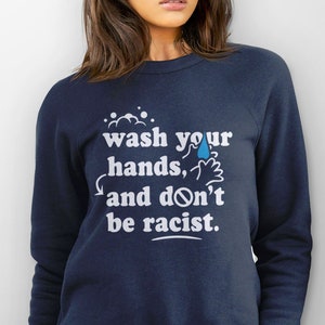 May include: A navy blue sweatshirt with a white graphic that says "wash your hands, and don't be racist." The graphic also includes a blue water drop and a hand washing symbol.