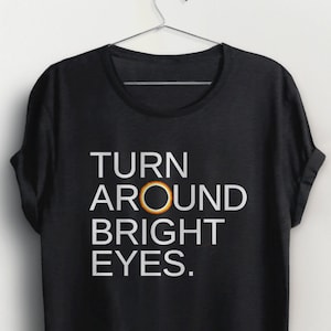 May include: Black t-shirt with white text that reads "TURN AROUND BRIGHT EYES." A gold ring is centred between the words "AROUND" and "BRIGHT".