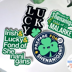Puede incluir: Cuatro pegatinas verdes y negras con frases de tem&aacute;tica irlandesa. Las pegatinas presentan tr&eacute;boles, las palabras "Lucky", "Irish", "Shenanigans" y "Malarkey". Una quinta pegatina presenta un tr&eacute;bol de dibujos animados con una cara sonriente y las palabras "&iexcl;Prep&aacute;rate para las travesuras!" Una peque&ntilde;a pegatina con un pastel y las palabras "Pegatinas resistentes a la intemperie e impermeables" se encuentra en la esquina inferior derecha.