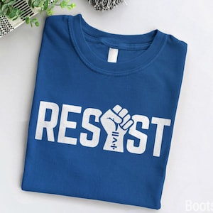 Könnte beinhalten: Ein königsblaues T-Shirt mit dem Wort "RESIST" in Weiß gedruckt, in der Mitte des Wortes befindet sich eine Grafik mit einer erhobenen Faust.