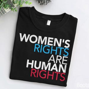 Könnte beinhalten: Schwarzes T-Shirt mit dem Text "Women's Rights Are Human Rights" in weißer, blauer und roter Schrift.