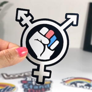 May include: A black and white sticker with a raised fist in the center. The fist is colored red, white, and blue. The fist is surrounded by a circle with a transgender symbol on either side.