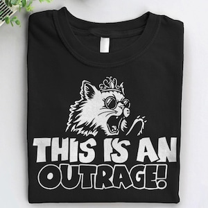 May include: Black t-shirt with a white graphic of a cat wearing sunglasses and a crown, with the text "THIS IS AN OUTRAGE!". The cat is roaring with its claws out. The shirt is folded neatly.
