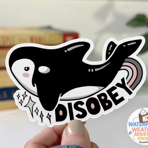 May include: A white sticker with a black and white cartoon orca swimming with a rainbow tail. The orca has a pink blush and is swimming towards the text "DISOBEY". The sticker has a sparkly design around the orca.