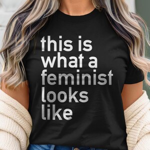 Puede incluir: Una camiseta negra con el texto "this is what a feminist looks like" impreso en blanco.