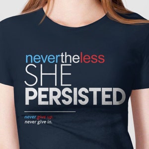 Puede incluir: Una camiseta azul marino con el texto "nevertheless she persisted" en blanco, con las palabras "never" en rojo y "less" en azul. El texto "never give up. never give in." est&aacute; impreso en blanco m&aacute;s peque&ntilde;o debajo del texto principal.