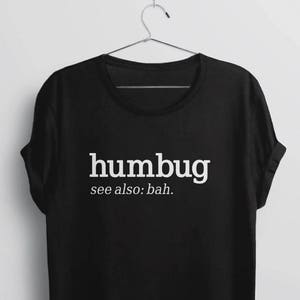 K&ouml;nnte beinhalten: Schwarzes T-Shirt mit dem wei&szlig;en Wort "humbug" und dem Zusatz "see also: bah.". Das T-Shirt h&auml;ngt an einem Metallb&uuml;gel. Die &Auml;rmel des T-Shirts sind hochgekrempelt.