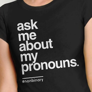 May include: Black t-shirt with white text that reads "ask me about my pronouns."  The text is in a bold, sans-serif font.  The hashtag "#nonbinary" is printed below the text.
