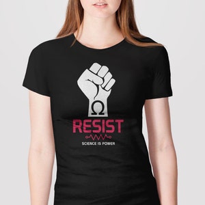 Pode incluir: Camiseta preta com um gr&aacute;fico de punho levantado branco e o texto "RESIST" em vermelho. Abaixo do texto h&aacute; um s&iacute;mbolo de resistor estilizado e o texto "SCIENCE IS POWER".