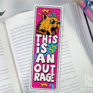 Puede incluir: Un marcap&aacute;ginas rosa con el texto "THIS IS AN OUTRAGE" y un gato de dibujos animados con gafas de sol y corona. El gato es naranja y amarillo con contorno negro. El marcap&aacute;ginas est&aacute; sobre un libro abierto.