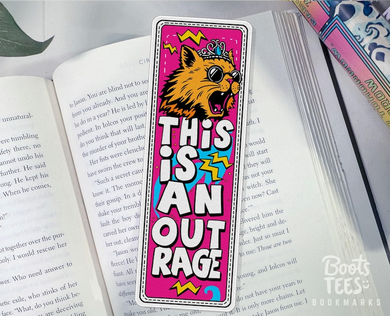 Puede incluir: Un marcap&aacute;ginas rosa con el texto "THIS IS AN OUTRAGE" y un gato de dibujos animados con gafas de sol y corona. El gato es naranja y amarillo con contorno negro. El marcap&aacute;ginas est&aacute; sobre un libro abierto.