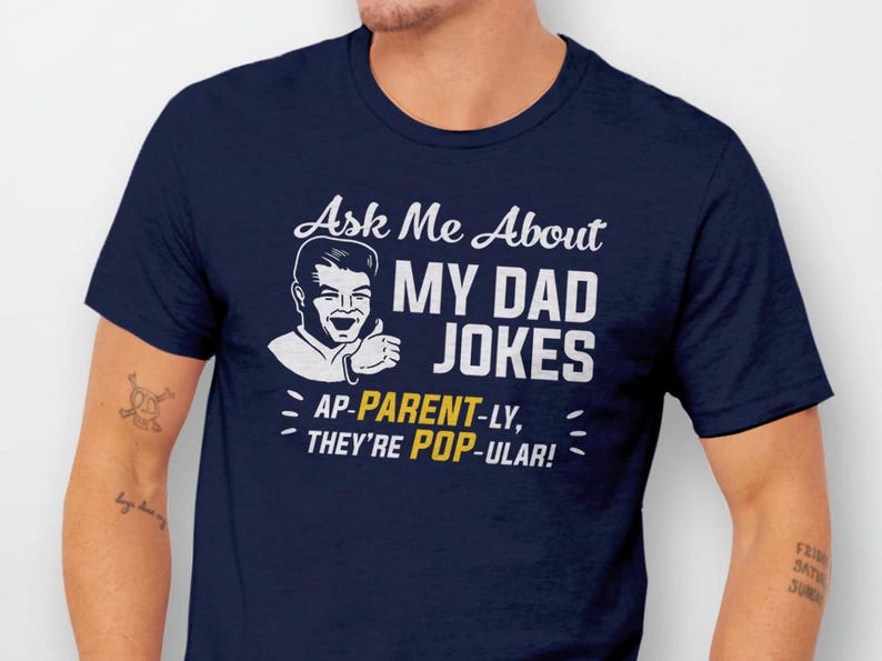 May include: Navy blue t-shirt with a white cartoon illustration of a man with the text "Ask Me About My Dad Jokes AP-PARENT-LY, THEY'RE POP-ULAR!"