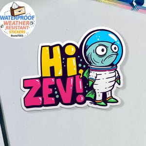 May include: A sticker featuring a cartoon alien astronaut with a blue helmet and a grumpy expression. The sticker says "Hi Zev!" in yellow and pink bubble letters. The sticker is waterproof and weather resistant.