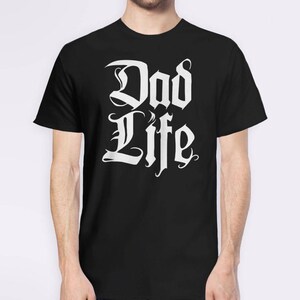 Puede incluir: Camiseta negra con texto blanco que dice "Dad Life" en una fuente estilizada.