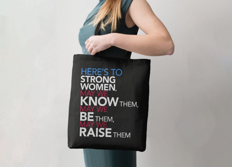 May include: Black tote bag with the text "HERE'S TO STRONG WOMEN, MAY WE KNOW THEM, MAY WE BE THEM, MAY WE RAISE THEM" in blue, pink, and white.