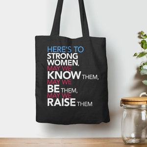 May include: Black tote bag with white text that reads "HERE'S TO STRONG WOMEN, MAY WE KNOW THEM, MAY WE BE THEM, MAY WE RAISE THEM"
