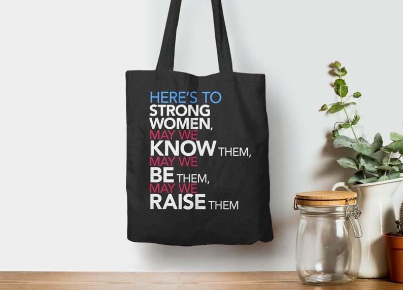 May include: Black tote bag with white text that reads "HERE'S TO STRONG WOMEN, MAY WE KNOW THEM, MAY WE BE THEM, MAY WE RAISE THEM"