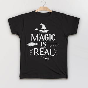 May include: Black t-shirt with a white graphic design featuring a witch's hat, a broom, and the text "Magic is Real".