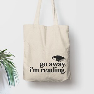 Puede incluir: Bolso tote de color crema con texto negro e ilustraci&oacute;n de libro. El texto dice "go away. I'm reading." El libro es un rect&aacute;ngulo negro simple con p&aacute;ginas blancas. El bolso tiene asas largas.