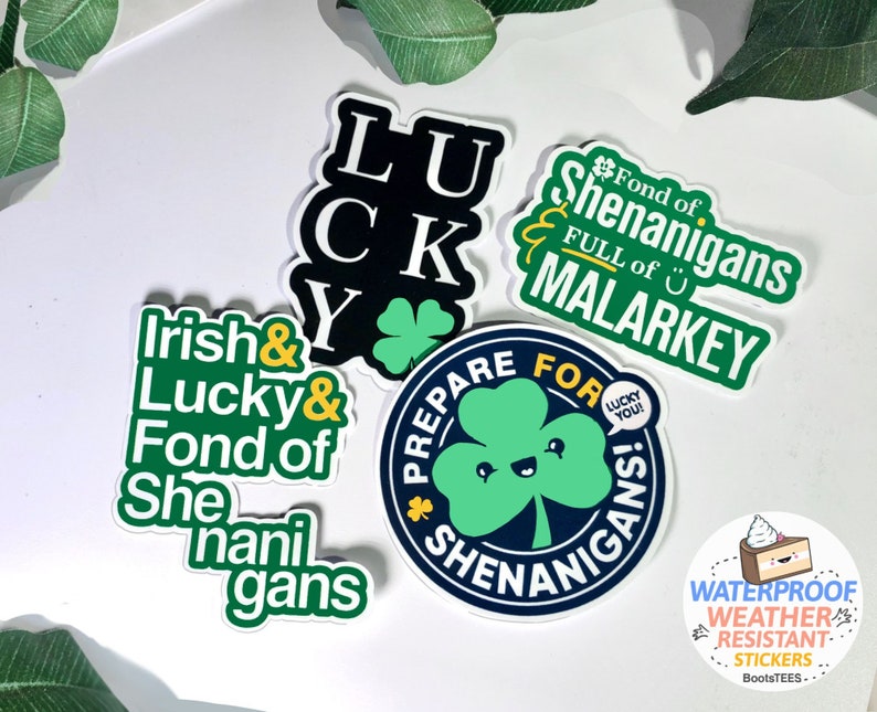 Puede incluir: Cuatro pegatinas verdes y negras con frases de tem&aacute;tica irlandesa. Las pegatinas presentan tr&eacute;boles, las palabras "Lucky", "Irish", "Shenanigans" y "Malarkey". Una quinta pegatina presenta un tr&eacute;bol de dibujos animados con una cara sonriente y las palabras "&iexcl;Prep&aacute;rate para las travesuras!" Una peque&ntilde;a pegatina con un pastel y las palabras "Pegatinas resistentes a la intemperie e impermeables" se encuentra en la esquina inferior derecha.