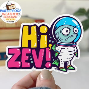 May include: A colorful sticker featuring a cartoon alien astronaut with a grumpy expression. The sticker says "Hi Zev!" in large, bold letters. The sticker is waterproof and weather resistant.