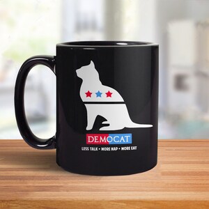 May include: Black coffee mug featuring a white silhouette of a cat wearing a red, white and blue collar with stars. The text "DEMOCAT" is below the cat and the text "LESS TALK - MORE NAP - MORE EAT" is below that.