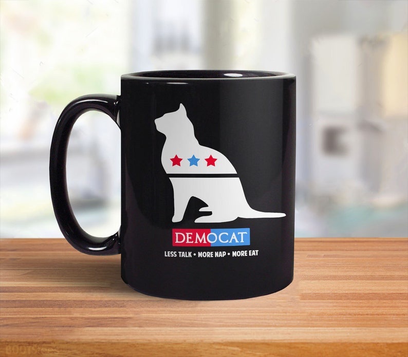 May include: Black coffee mug featuring a white silhouette of a cat wearing a red, white and blue collar with stars. The text "DEMOCAT" is below the cat and the text "LESS TALK - MORE NAP - MORE EAT" is below that.