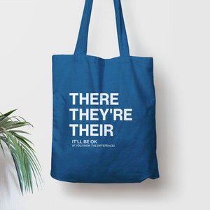 Op de afbeelding: Een blauwe tote bag met de woorden "THERE THEY'RE THEIR" in wit gedrukt. Onder de woorden staat de tekst "IT'LL BE OK IF YOU KNOW THE DIFFERENCE."