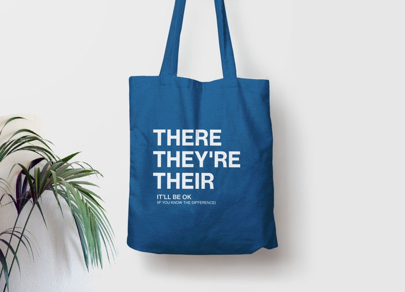 Op de afbeelding: Een blauwe tote bag met de woorden "THERE THEY'RE THEIR" in wit gedrukt. Onder de woorden staat de tekst "IT'LL BE OK IF YOU KNOW THE DIFFERENCE."
