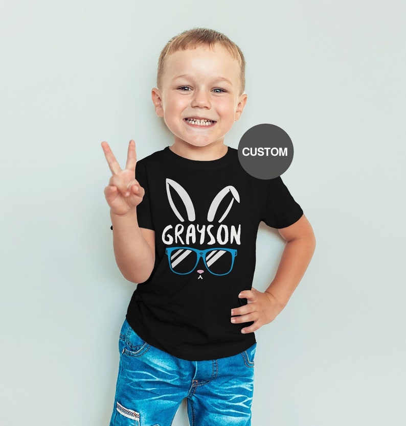 May include: Black t-shirt with a white bunny rabbit graphic wearing blue sunglasses and the name "GRAYSON" printed on the front. The shirt is being worn by a young child.