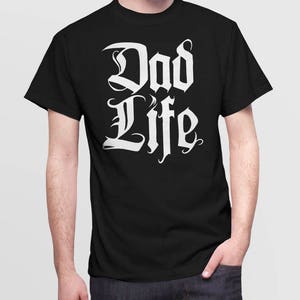 Puede incluir: Camiseta negra con texto blanco que dice "Dad Life" en una fuente estilizada.