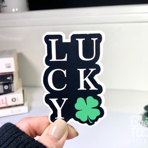 Puede incluir: Una pegatina negra y blanca con la palabra "LUCKY" escrita verticalmente con un tr&eacute;bol de cuatro hojas verde en la parte inferior.