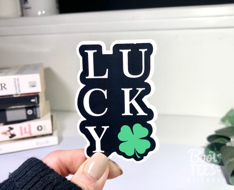 Puede incluir: Una pegatina negra y blanca con la palabra "LUCKY" escrita verticalmente con un tr&eacute;bol de cuatro hojas verde en la parte inferior.