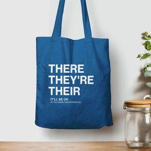 Op de afbeelding: Een blauwe tote bag met de woorden "THERE THEY'RE THEIR" in wit gedrukt. Onder de woorden staat de tekst "IT'LL BE OK IF YOU KNOW THE DIFFERENCE".
