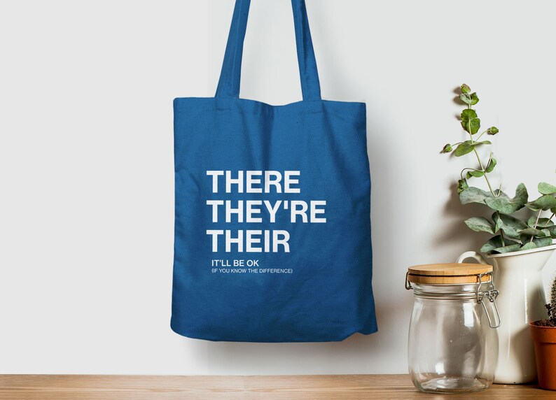 Op de afbeelding: Een blauwe tote bag met de woorden "THERE THEY'RE THEIR" in wit gedrukt. Onder de woorden staat de tekst "IT'LL BE OK IF YOU KNOW THE DIFFERENCE".