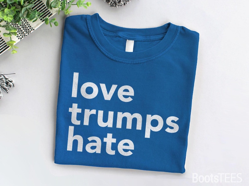 Puede incluir: Una camiseta azul con el texto "love trumps hate" impreso en blanco.