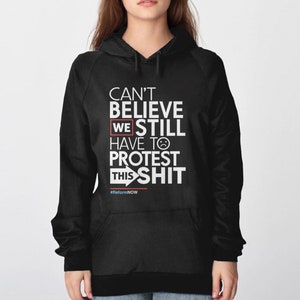 May include: Black hooded sweatshirt with white text that reads "CAN'T BELIEVE WE STILL HAVE TO PROTEST THIS SHIT." The text includes a sad face emoji and the hashtag #ReformNOW.
