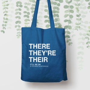 Op de afbeelding: Een blauwe tote bag met de woorden "THERE THEY'RE THEIR" in wit gedrukt. Onder de woorden staat de tekst "IT'LL BE OK (IF YOU KNOW THE DIFFERENCE)".