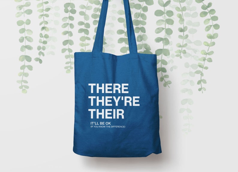 Op de afbeelding: Een blauwe tote bag met de woorden "THERE THEY'RE THEIR" in wit gedrukt. Onder de woorden staat de tekst "IT'LL BE OK (IF YOU KNOW THE DIFFERENCE)".