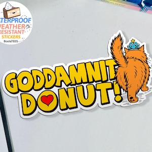May include: A sticker with the text "GODDAMNIT DONUT!" in yellow, outlined in black, with a red heart replacing the "O". An orange cat wearing a blue crown is on the right. The sticker is waterproof and weather resistant.