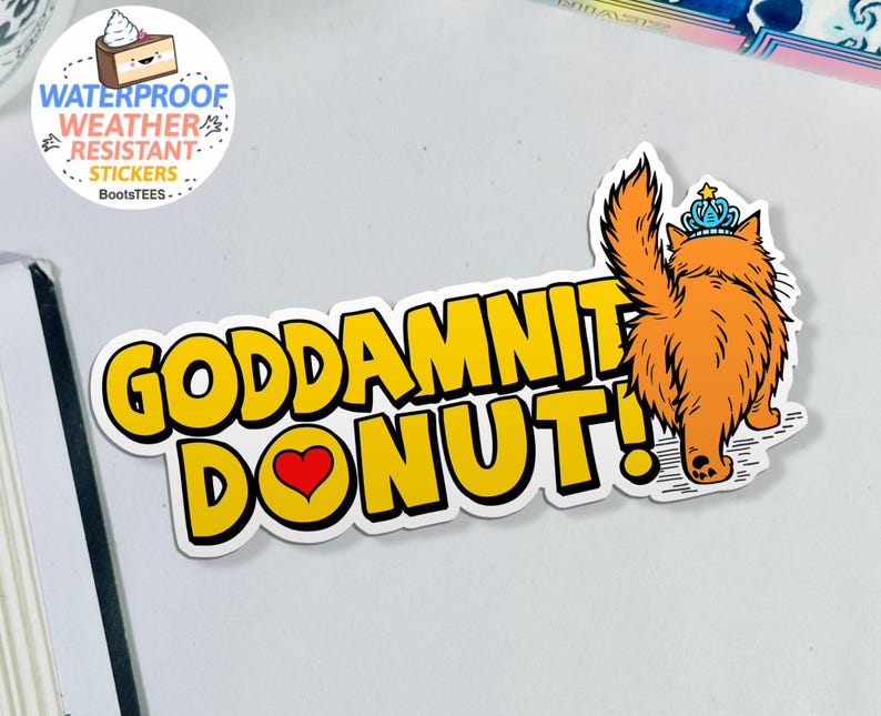 May include: A sticker with the text "GODDAMNIT DONUT!" in yellow, outlined in black, with a red heart replacing the "O". An orange cat wearing a blue crown is on the right. The sticker is waterproof and weather resistant.