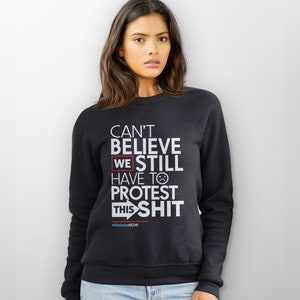 May include: Black sweatshirt with white text that reads "CAN'T BELIEVE WE STILL HAVE TO PROTEST THIS SHIT." The word "WE" is highlighted in a red box. Below the text is the hashtag "#ReformNOW".
