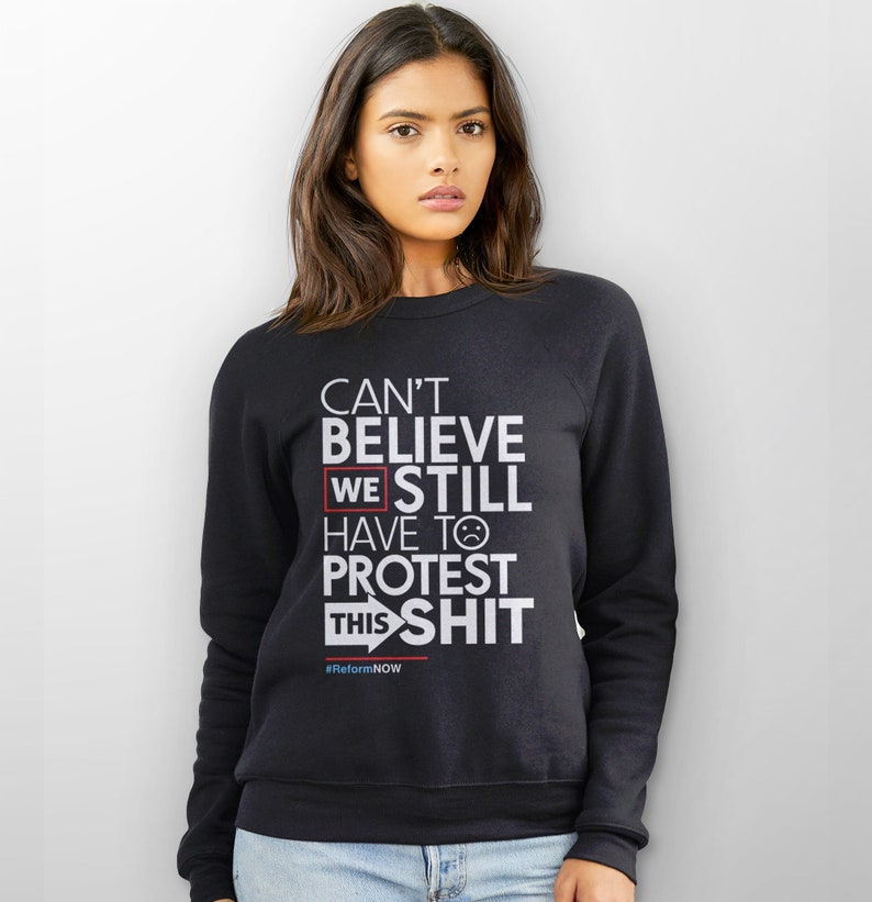 May include: Black sweatshirt with white text that reads "CAN'T BELIEVE WE STILL HAVE TO PROTEST THIS SHIT." The word "WE" is highlighted in a red box. Below the text is the hashtag "#ReformNOW".