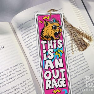 Puede incluir: Un marcap&aacute;ginas colorido con un gato con gafas de sol y una corona, con el texto "THIS IS AN OUTRAGE". El marcap&aacute;ginas tiene una borla dorada y est&aacute; colocado en un libro abierto.