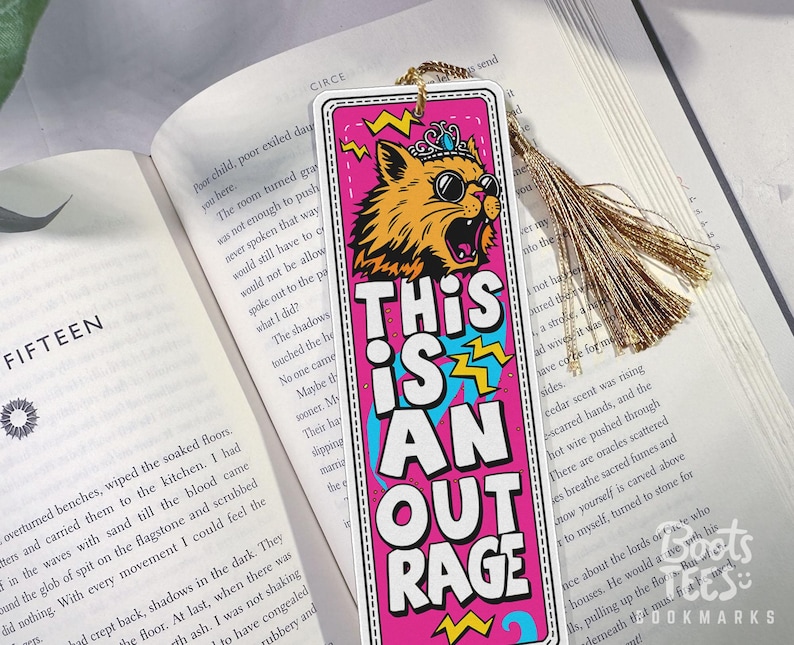 Puede incluir: Un marcap&aacute;ginas colorido con un gato con gafas de sol y una corona, con el texto "THIS IS AN OUTRAGE". El marcap&aacute;ginas tiene una borla dorada y est&aacute; colocado en un libro abierto.