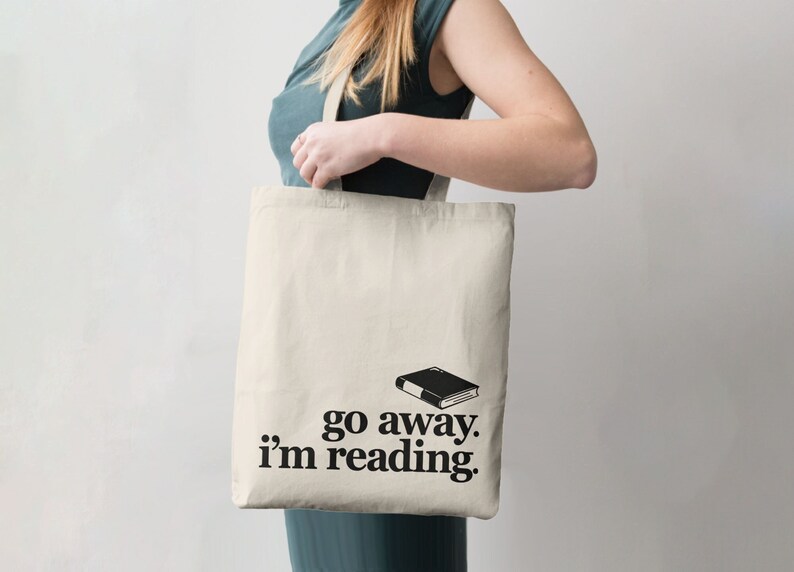 Puede incluir: Bolso tote de color crema con el texto "go away. i'm reading." y una ilustraci&oacute;n de libro negro. El bolso lo sostiene una persona. El bolso est&aacute; hecho de una tela resistente.