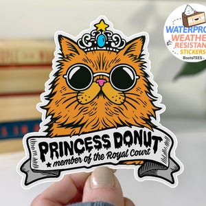 May include: A sticker featuring an orange cat wearing sunglasses and a crown. The cat has the words "PRINCESS DONUT" on a banner, with "member of the Royal Court" below. The sticker is waterproof and weather-resistant.