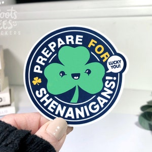 Puede incluir: Una pegatina redonda con fondo azul y borde blanco. La pegatina presenta un tr&eacute;bol verde con una cara sonriente y el texto "Prepare for Shenanigans!" en blanco. Una burbuja de di&aacute;logo amarilla con el texto "Lucky You!" est&aacute; al lado del tr&eacute;bol.